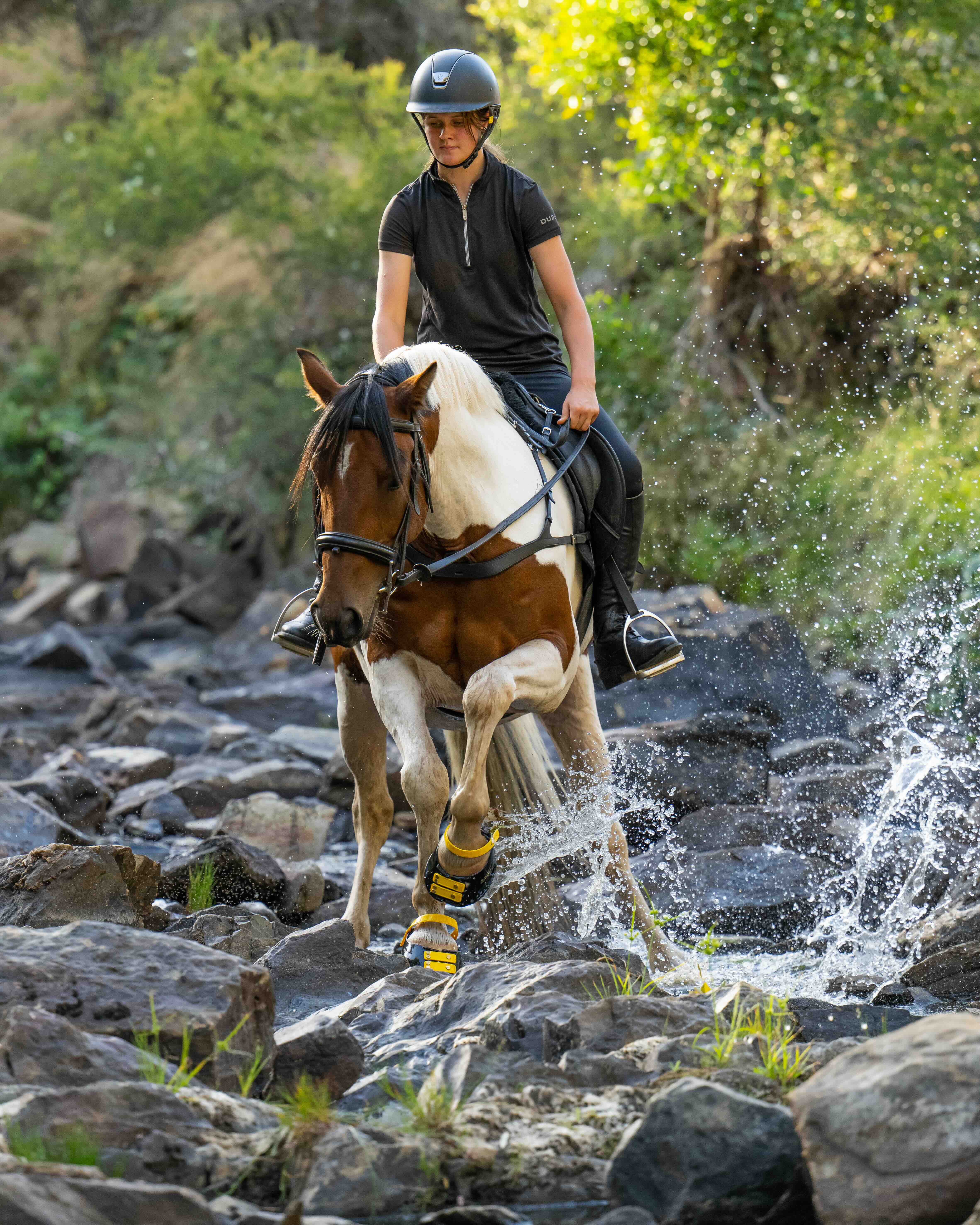 Een ruiter op een bruin-wit gevlekt paard dat door een rotsachtige rivier waadt; het paard draagt geel met zwarte hoefschoenen voor optimale grip en bescherming.